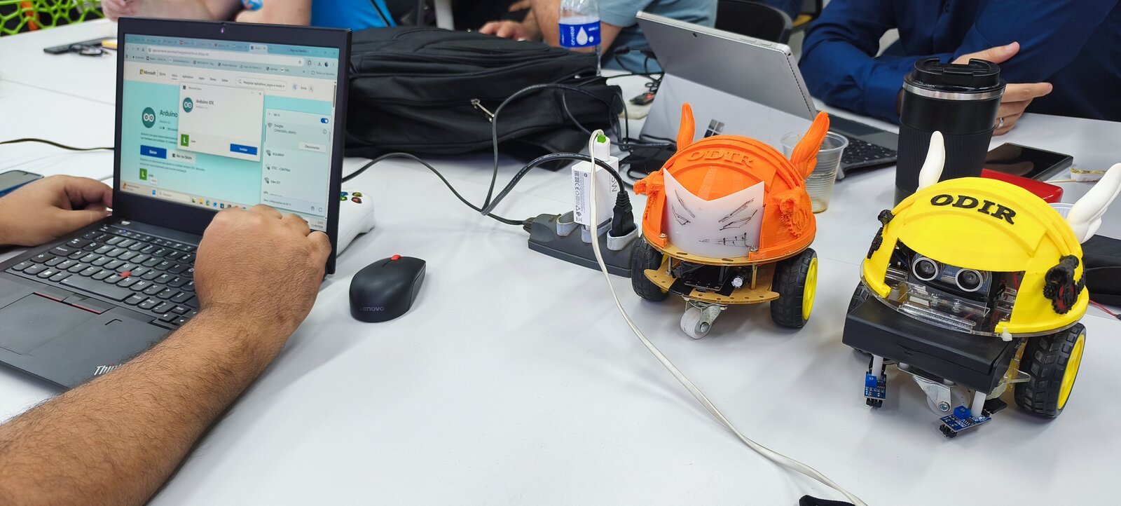 Oficina de robótica com ODIR laranja e amarelo sobre a mesa, ao lado de um notebook com a IDE do Arduino