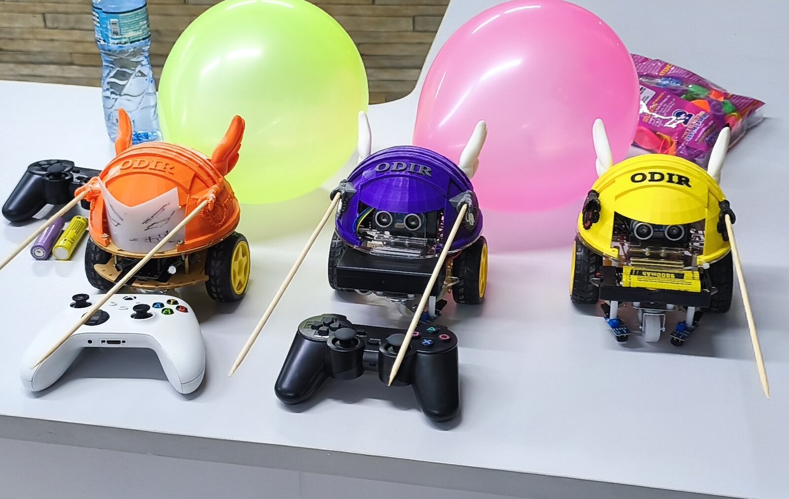 Três ODIR nas cores laranja, roxo e amarelo, prontos para o Duelo de Bexigas com controles de videogame